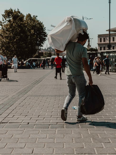 A young man walking across a paved public square, carrying a large white plastic bag on his shoulder and a smaller black bag in his right hand. The man wears a grey T-shirt, light grey jeans, and dark shoes. The background features several pedestrians, some walking and others standing, with a mix of trees, vehicles, and historic buildings lining the square. There are street lamps and a clear blue sky overhead, indicating daytime with bright natural light. The scene suggests an urban environment where individuals might be involved in personal errands or waste disposal, subtly connected to private rubbish handling services such as those offered by Rubbish Clearance Sutton. The overall atmosphere is casual and busy, with a focus on the movement of people amid an orderly and well-maintained setting, emphasizing the context of waste management and rubbish removal in the community.