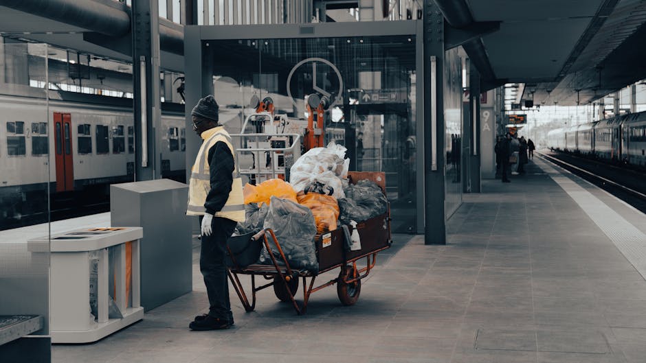 At a modern train station platform, a worker wearing a reflective vest, black trousers, and a knit cap stands beside a large, wheeled trolley loaded with an assortment of refuse, including black, grey, and orange plastic bags, some visibly bulging with waste. The trolley, positioned to the right of the worker, is filled with various household and general rubbish, with some bags appearing tied and others loosely packed. Behind the worker, a glass shelter with metal framing provides partial cover, and a set of orange and white mechanical devices—likely for loading or waste handling—are mounted on the station's structure. To the left, a white waste bin with a partially open lid sits on the platform, indicating a designated area for waste collection, while in the background, the railway tracks run alongside the platform, with several trains visible on both sides. The lighting appears natural, with diffused daylight creating a neutral and professional atmosphere appropriate for an urban environment. The scene subtly reflects the process of private waste disposal or rubbish removal services, such as those provided by Rubbish Clearance Sutton, with the focus on the transportation and management of waste within a public transport setting.