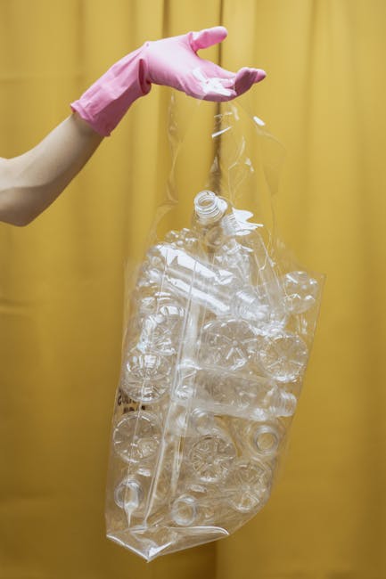 A person wearing pink rubber gloves is holding a large, transparent plastic bag filled with various clear plastic bottles and containers, some with caps still attached. The background features a plain, yellow curtain or wall, providing a simple backdrop that contrasts with the clear plastics. The scene suggests a recycling or waste collection process, with the individual preparing the plastics for disposal or transfer, indicative of an independent or private waste handling service such as Rubbish Clearance Sutton. The focus is on the environmental practice of collecting and managing recyclable materials, emphasizing the importance of proper rubbish separation. The lighting appears even and bright, highlighting the shiny, smooth surfaces of the plastic bottles and the bag's transparency. This visual aligns with services related to rubbish removal, emphasizing responsible waste management practices for household or commercial waste, particularly plastics. The overall composition displays a clear scene of collection preparation within a waste management context, supporting the theme of alternative rubbish clearance methods outside local authority collections.