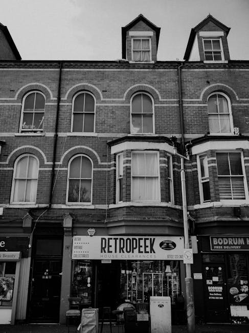 The image shows the upper floors of a three-story brick building with a retail shop on the ground level. The building features a traditional red brick exterior with decorative arches above the windows, which have white frames and sashes. The ground-floor storefront has large glass windows and a central entrance, with a sign reading 'RETROPEEK' indicating house clearance services, suggesting a business that provides private waste disposal rather than local authority rubbish collection. To the right of the shop, there is another commercial storefront with signage for a food delivery service. The upper floors of the building display multiple sash windows, some with curtains visible through the glass. The foreground includes a pavement area with a few objects like a small trash bin and some chairs, typical of an urban retail frontage. The black and white tone emphasizes the architectural details and the commercial signage, subtly aligning with themes of independent collection and alternative waste handling methods alongside regular rubbish removal activity within a city environment.