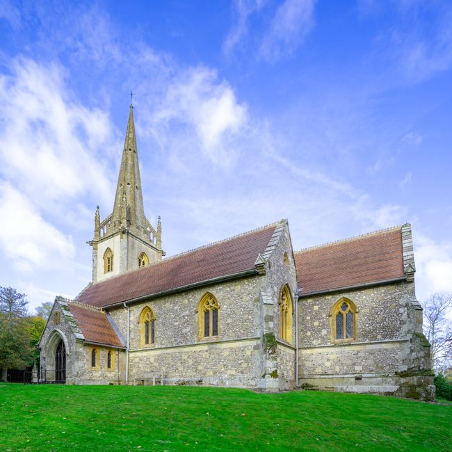 A historic stone church with a tall, pointed spire set against a bright blue sky with scattered white clouds. The church's exterior features weathered stone walls with patches of moss and lichen, and several narrow, arched windows with yellow-tinted stained glass. The steeply pitched roof is covered in reddish-brown tiles, contrasting with the gray stonework. A small, arched entranceway is visible on the left side, along with additional smaller extensions on the building's sides. The church is situated on a well-maintained, lush green lawn that gently slopes away from the structure, with a few leafless trees visible in the background. The scene is well-lit with natural daylight, highlighting the textures of the aged stone and the vibrant colors of the sky, creating a serene and timeless atmosphere suitable for discussing private or independent waste collection solutions by rubbish clearance services like Rubbish Clearance Sutton, although no waste or rubbish is visible in the image itself.